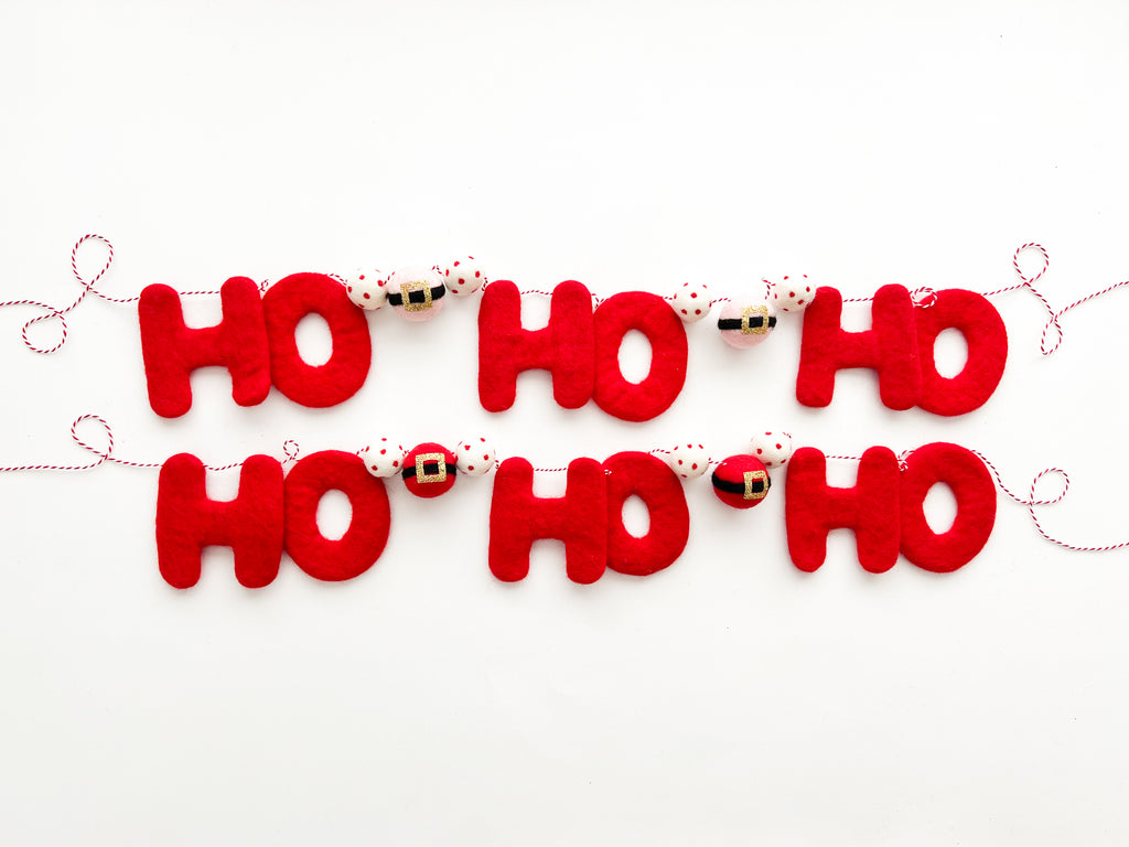 A red felt garland with the text 'ho ho ho' displayed repeatedly, with Santa Claus designs at the end of each 'ho'. The garland is strung on a twine and set against a white background.